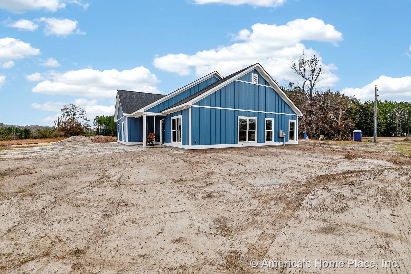 Blue vertical siding with white trim accents, large rectangular windows, covered entry porch, black shingle roof, unfinished yard, modern farmhouse single-story exterior.