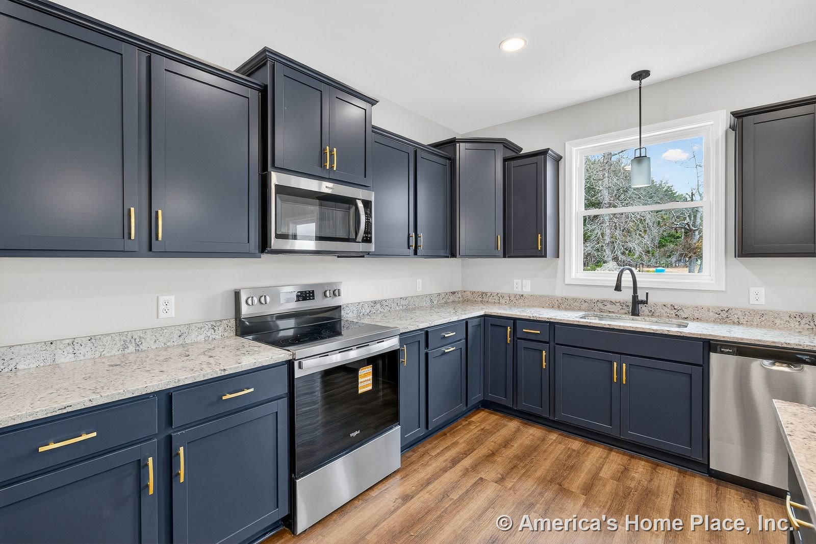 Navy shaker cabinets with gold hardware, granite countertops, stainless steel oven and microwave, large window above built-in sink, pendant light fixture, wood-look flooring