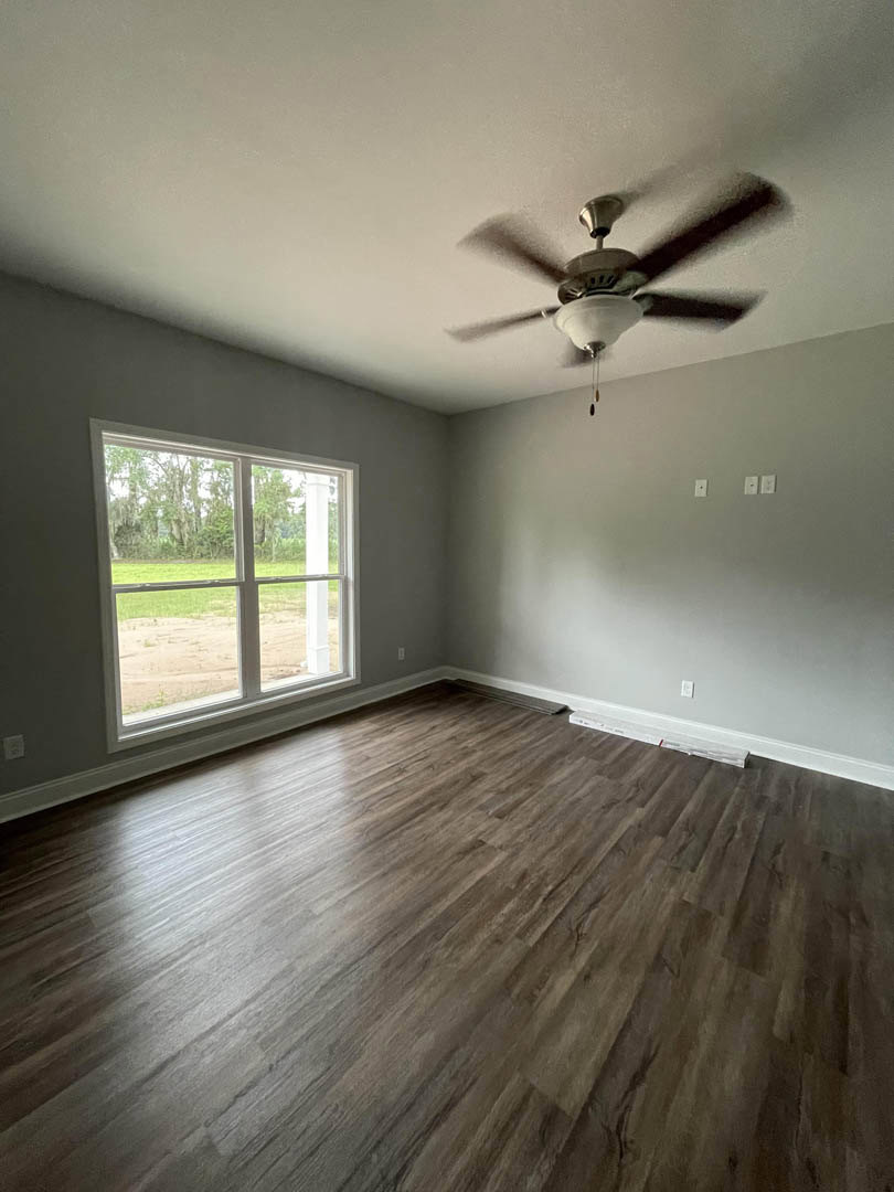Bedroom with wood flooring, white plaster walls, ceiling fan with light fixture, and large window overlooking grassy field
