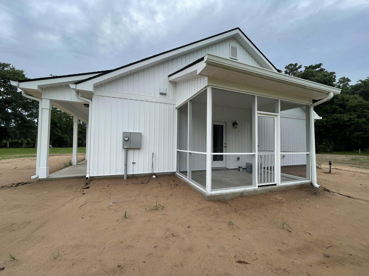 White siding house with covered porch, glass screen door, white-framed window, grey electrical box and pipe on exterior wall, shaded by nearby trees.