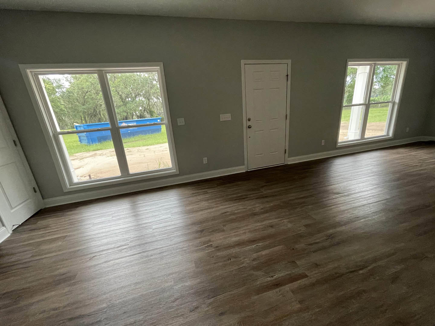 Bright room with laminate wood flooring, white walls, large windows with white frames showing trees outside, and a white door with a silver handle