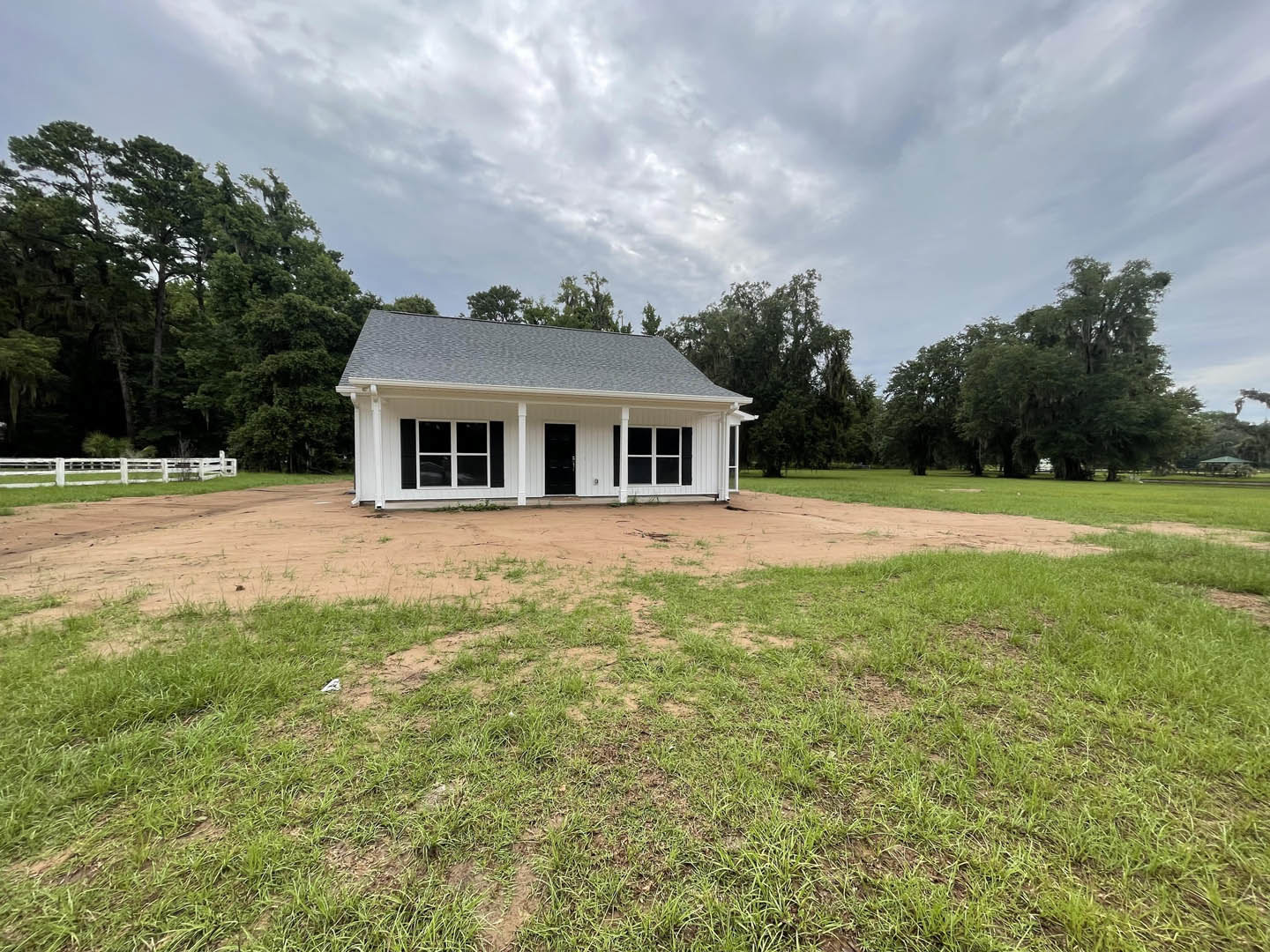 White house with black doors and white-framed windows, dirt driveway leading up to the entrance, surrounded by grassy field, white fence, and trees in the background under a partly
