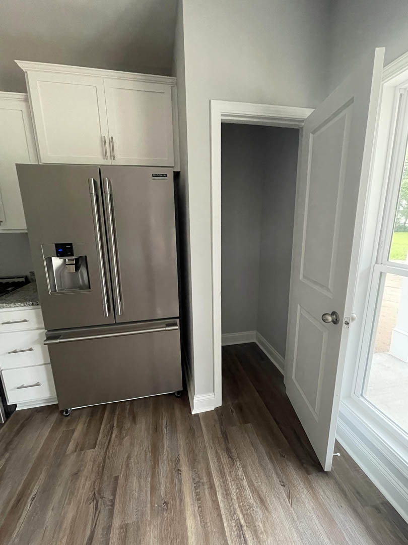 Modern kitchen featuring wood flooring, white cabinetry with silver handles, stainless steel refrigerator, open door, and a window with a white frame.