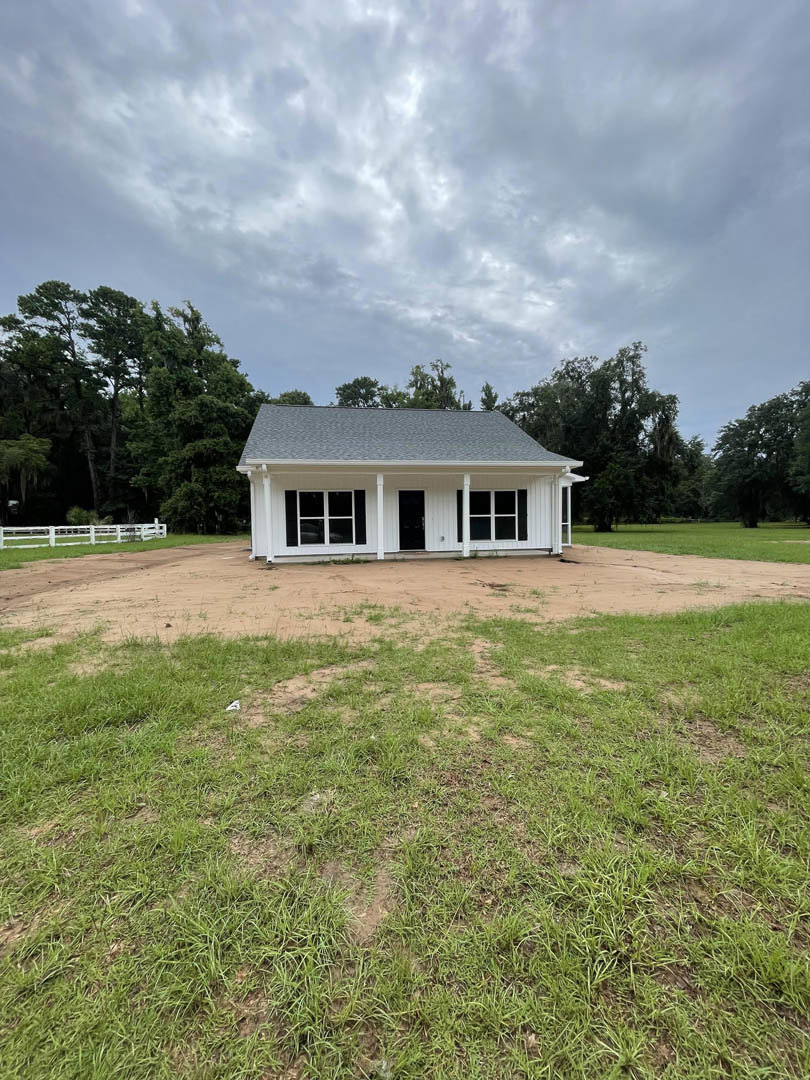White house with columned porch, wooden fence, dirt patch in front, surrounded by trees under cloudy sky