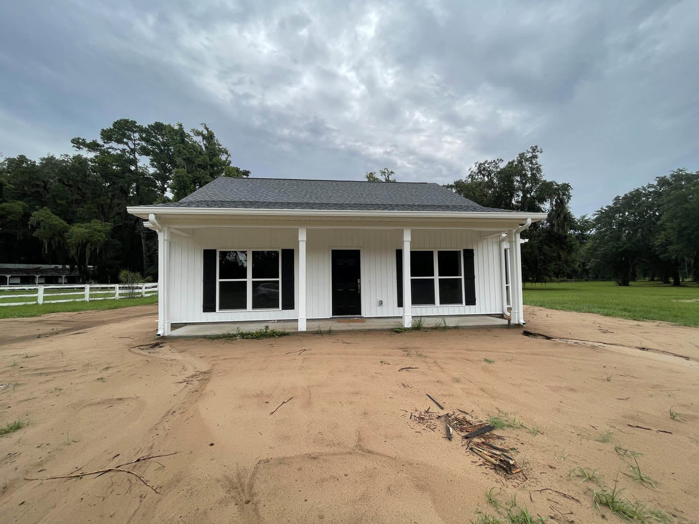 White siding house with black door and windows, surrounded by trees and a dirt road, under a cloudy sky, white fence visible in grassy field.