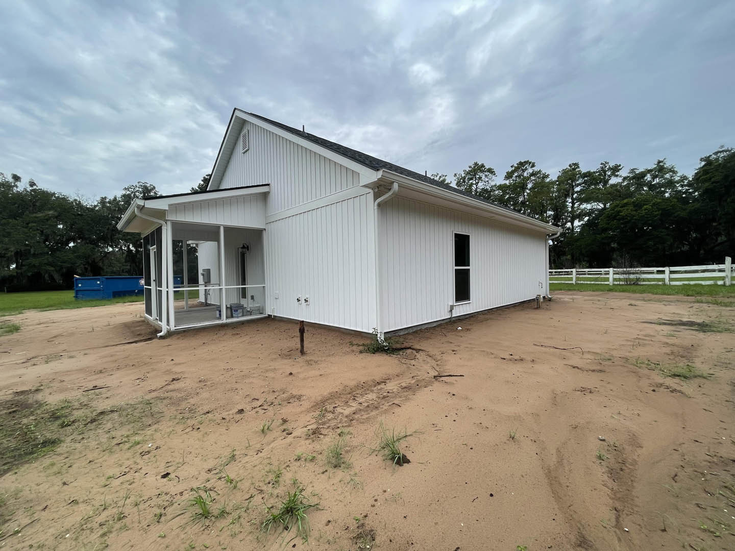 White house with gabled roof, white picket fence, dirt yard in foreground, blue container near entrance, sparse trees and cloudy sky in background