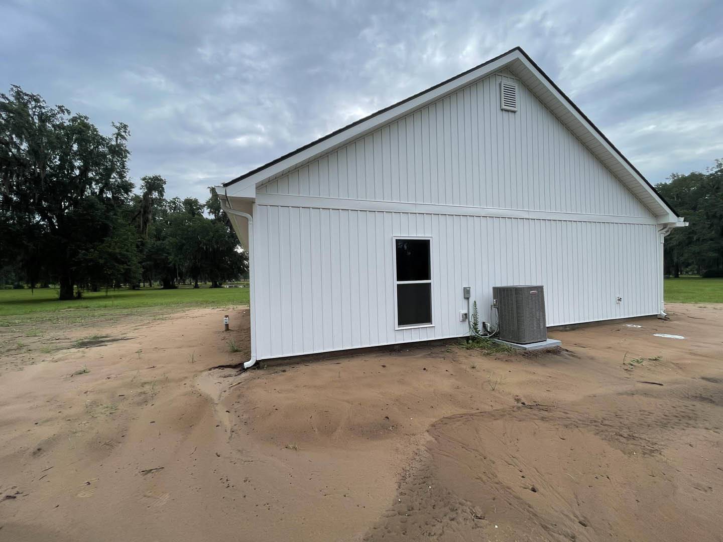 White modern house with large black-framed window, white door, grey heat pump unit, leafy tree, and bare dirt yard under cloudy sky