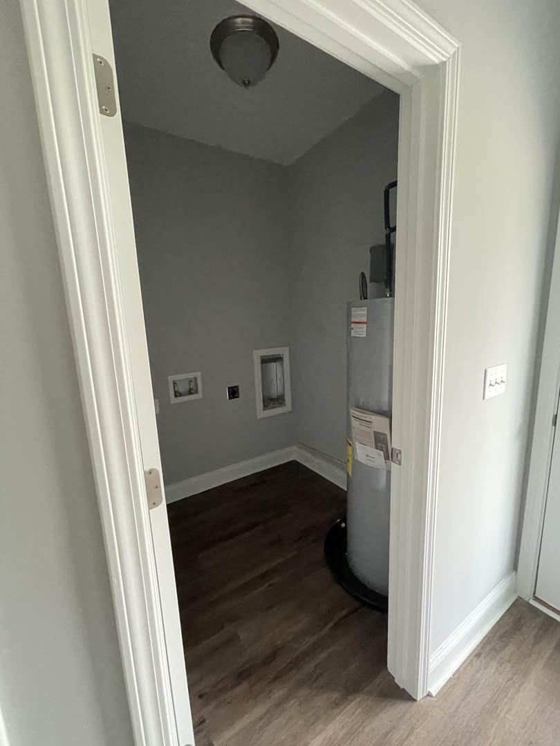 White paneled door opening into a bathroom with plaster walls, light wood flooring, and a white ceramic urinal visible near a metal fixture.