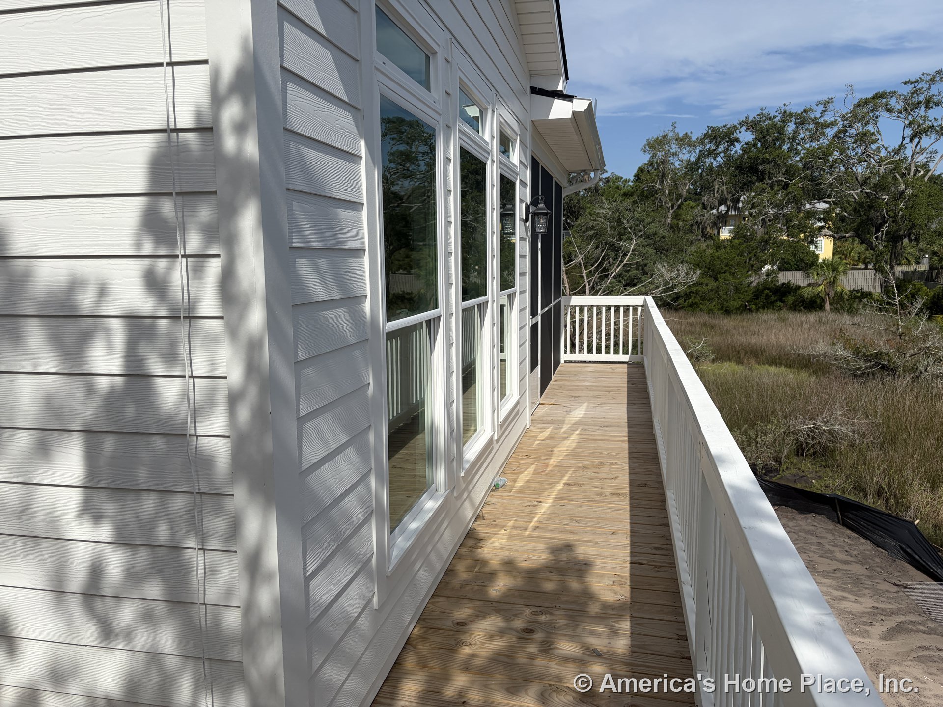 Wooden deck with white railing runs alongside home featuring white horizontal siding, expansive exterior windows, outdoor wall light fixture, and covered porch area.