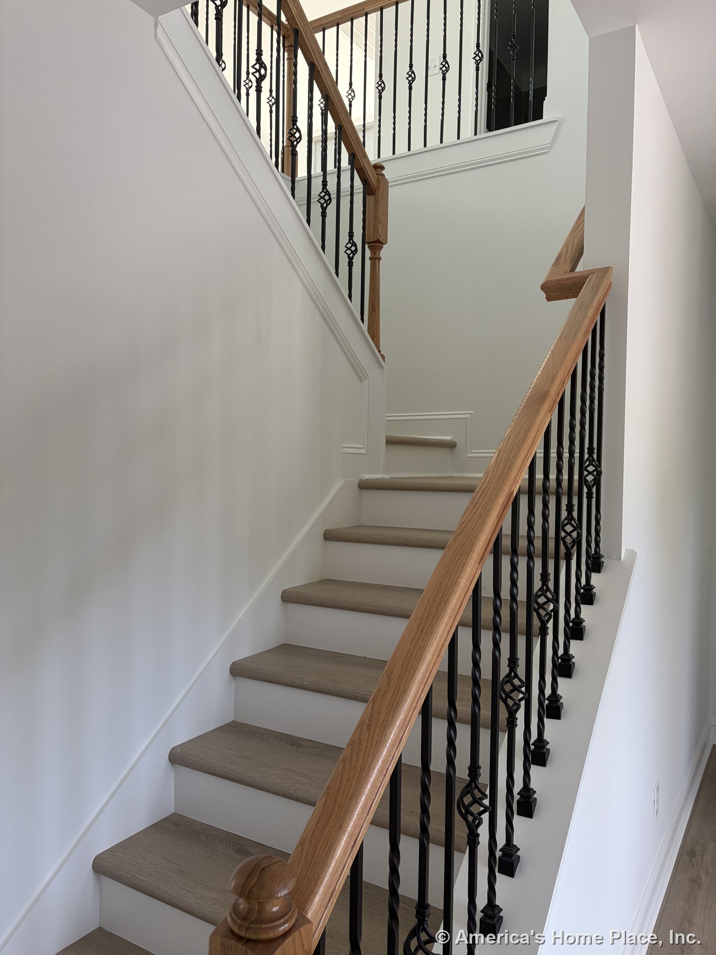 Staircase with light wood treads, white risers, black wrought iron balusters featuring decorative metalwork, wood handrail, wall trim detail, and upper landing with matching