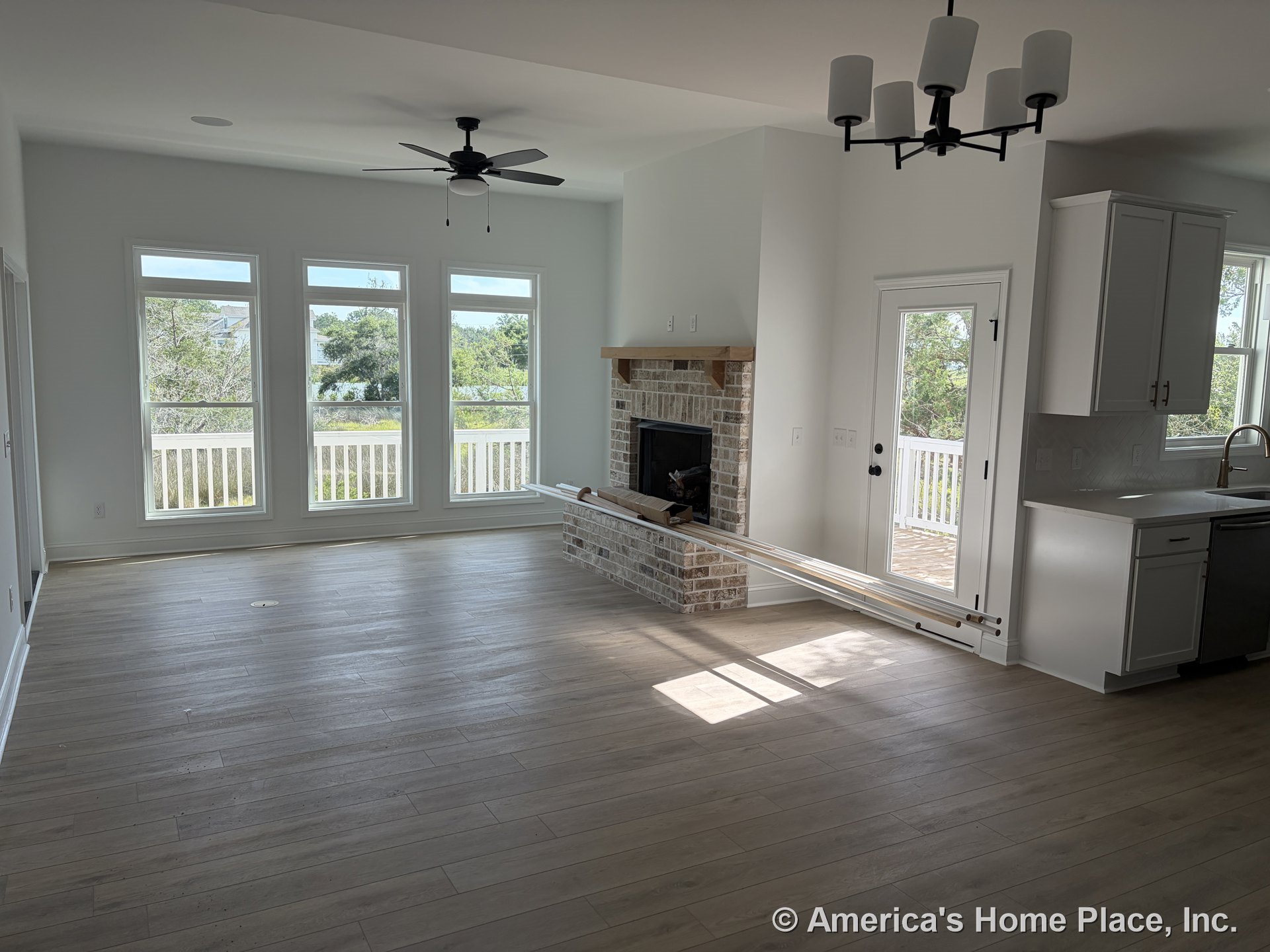 Open living area with three tall windows, brick fireplace with wood mantel, light wood plank flooring, black ceiling fan, modern chandelier, white kitchen cabinetry, and natural