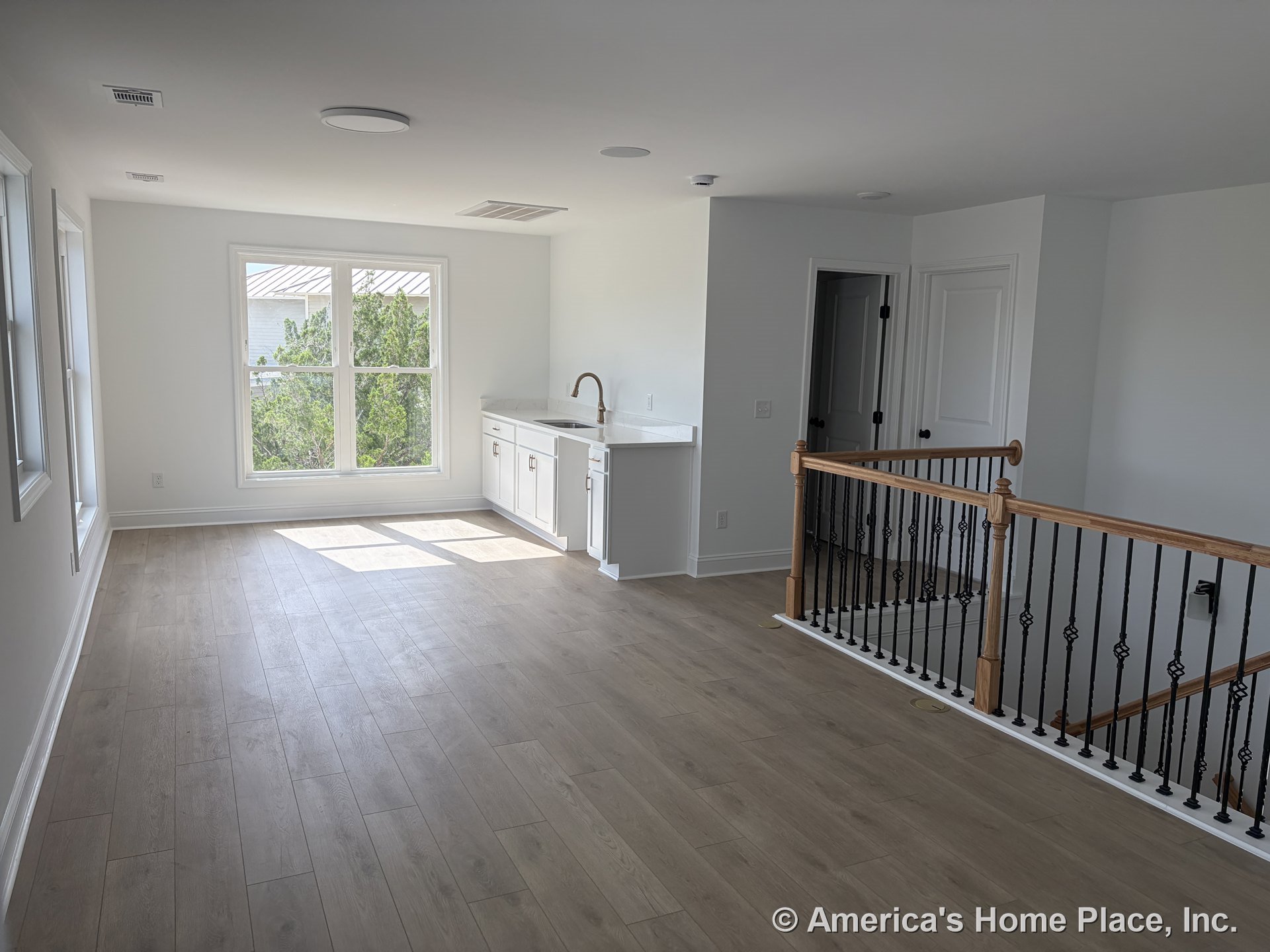 Upstairs loft with expansive double windows, light wood plank flooring, built-in wet bar featuring white cabinetry and white quartz countertop, black iron stair railing with iron