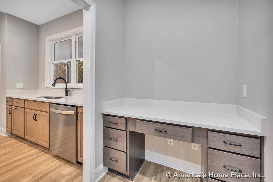 Light wood kitchen cabinets with white solid surface countertops, built-in desk area with drawers, stainless steel dishwasher, single-handle faucet, and double window above the