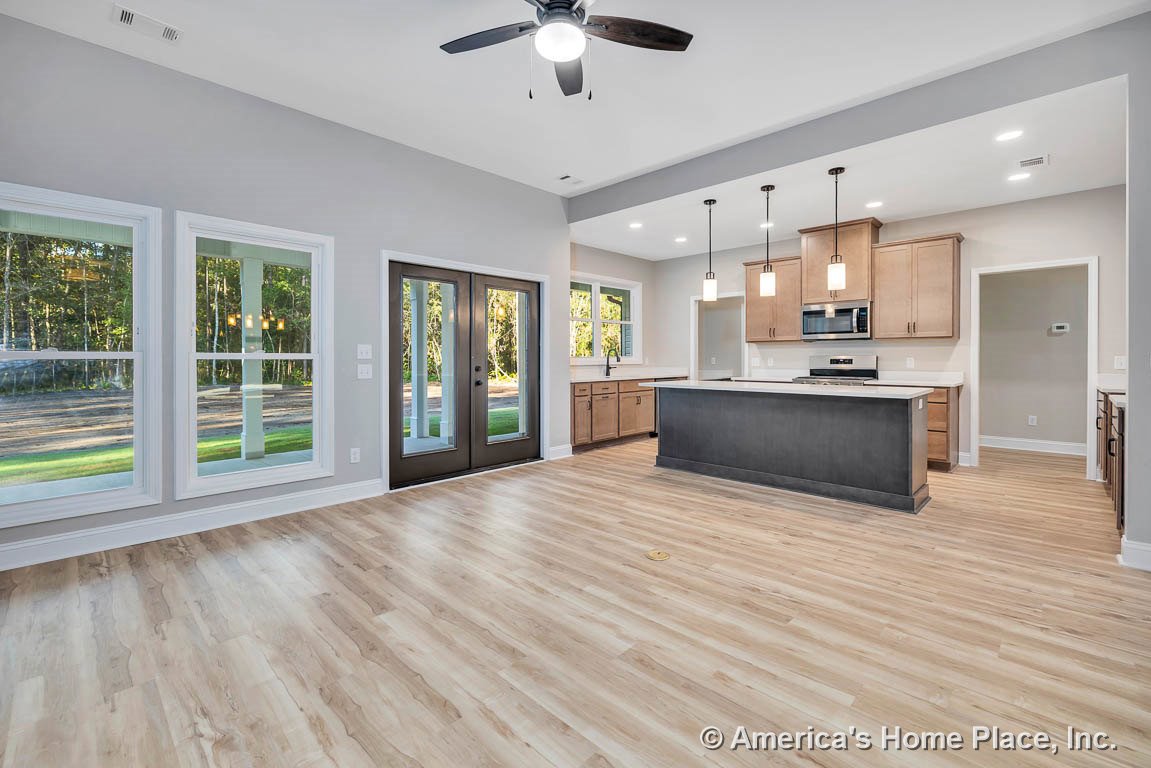 Open concept kitchen and living area with light wood plank flooring, large double windows, dark framed glass doors, central kitchen island topped with pendant lights, built-in