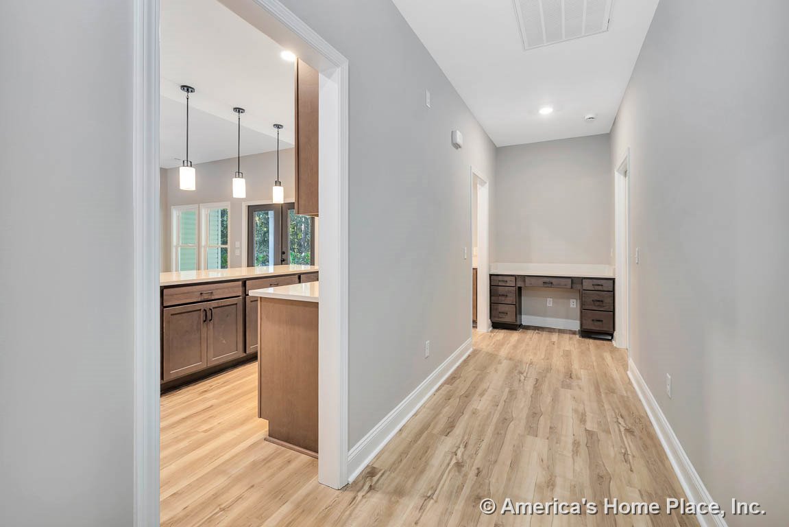 Light wood plank flooring in hallway with built-in desk cabinetry and drawers, recessed ceiling lighting, white trim and baseboards, view into kitchen featuring dark wood cabinets