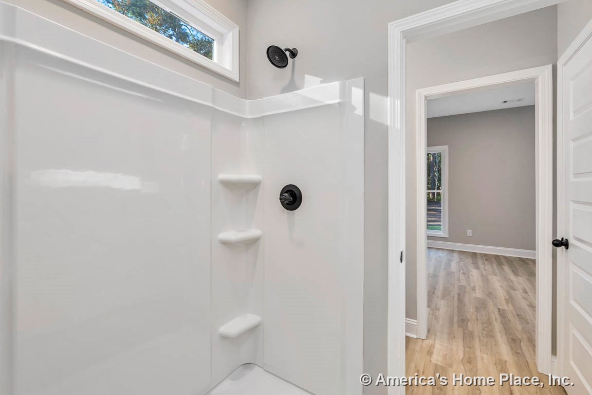 White fiberglass shower enclosure with built-in shelves, matte black showerhead and controls, transom window above shower, light wood flooring visible in adjacent room, neutral