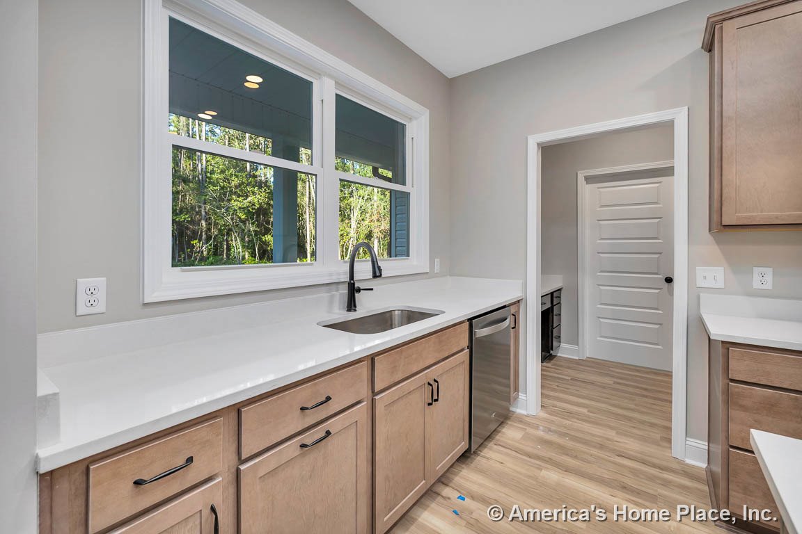 Light wood kitchen cabinets with white quartz countertops, stainless steel undermount sink set beneath double windows with white trim, built-in dishwasher, light wood flooring
