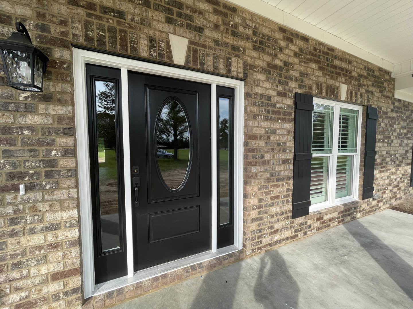 Black door with glass window set in red brick exterior, black lantern fixture mounted beside entrance, concrete ground visible.