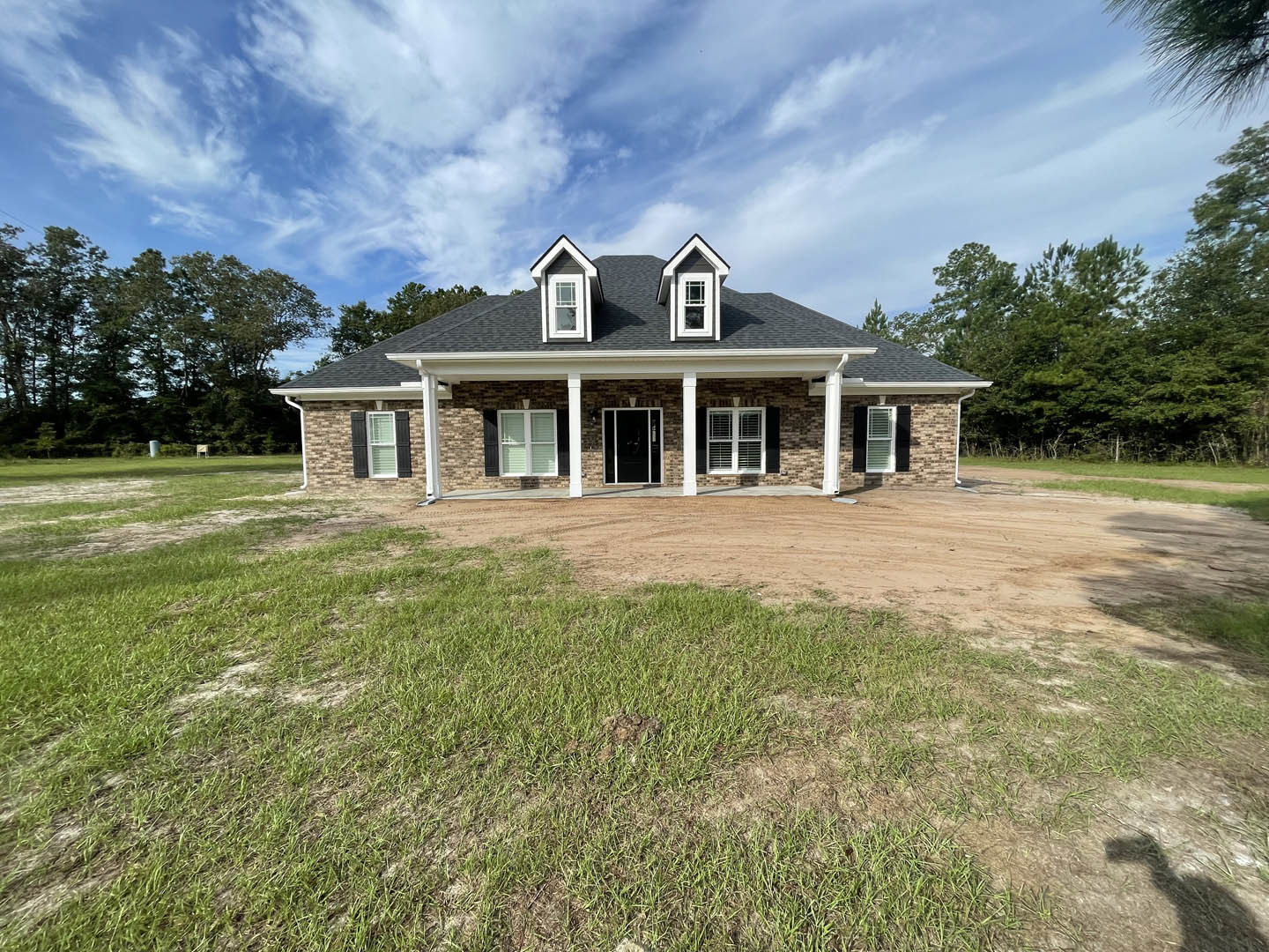 Two-story house with black front door, white-framed windows with blinds, glass door reflecting a person, dirt road bordered by grass, trees and cloudy sky in background