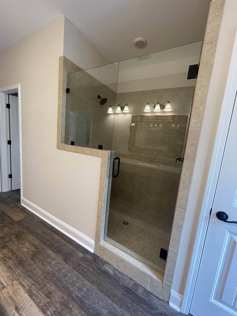 Glass shower door with chrome hinge, wood flooring adjacent to white tile shower, black handle on white vanity, close-up of modern lamp, plaster walls and ceiling