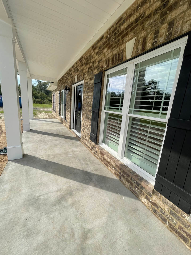 Red brick house with covered porch, white-trimmed window featuring black shutters, concrete sidewalk, and decorative front door