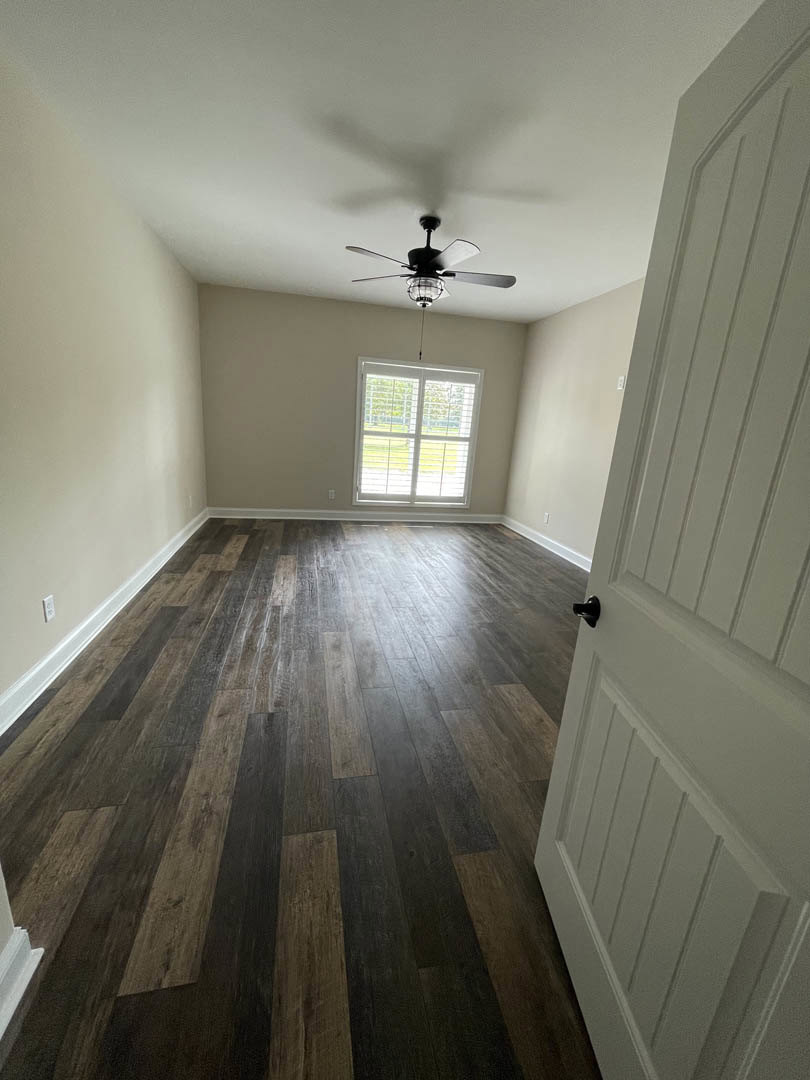 Ceiling fan with light fixture, hardwood floor reflecting natural light, white door with black handle, window with closed blinds, pale walls.