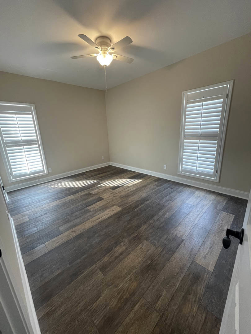 Ceiling fan with light fixture above hardwood floor, white walls, window with blinds, and door handle visible in custom home interior.