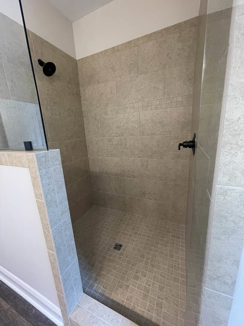 Glass shower enclosure with black soap dispenser mounted on white marble wall, black object on light tile floor, linear drain visible near shower entrance