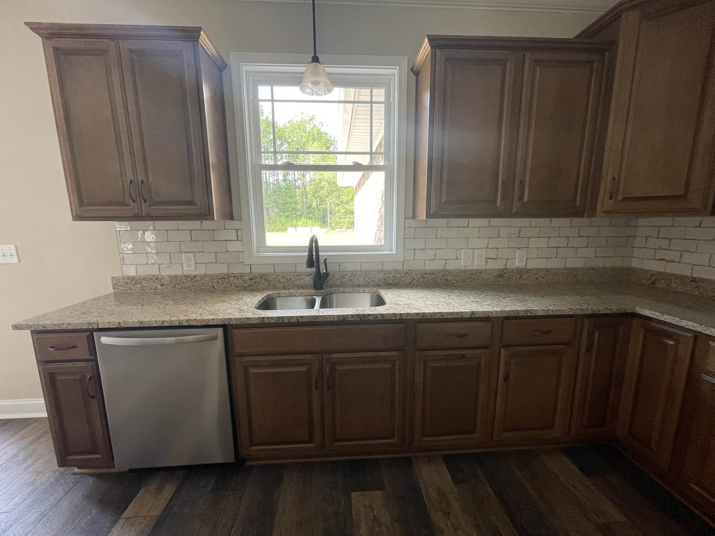 Granite countertop kitchen with stainless sink, white cabinetry, tile backsplash, built-in dishwasher, ceiling light fixture, and window overlooking trees.