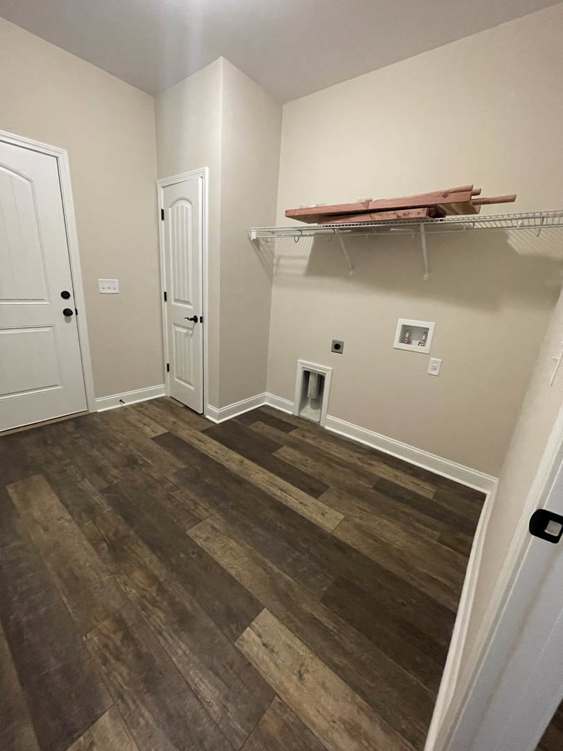 Wood flooring with white walls, built-in shelf, white door featuring black hardware, window with white trim, and silver fixtures in a bathroom interior.