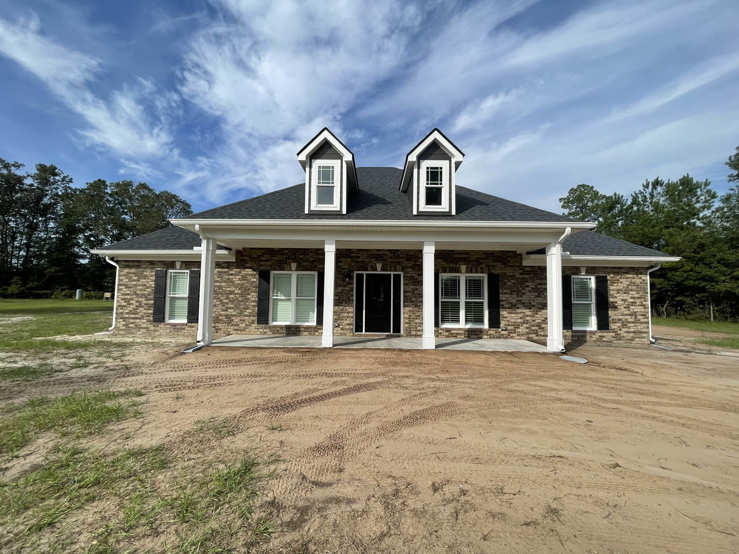 Black and white roofed house with glass door in white frame, windows with blinds and white trim, tire tracks on dirt road leading to porch, surrounded by trees under cloudy sky