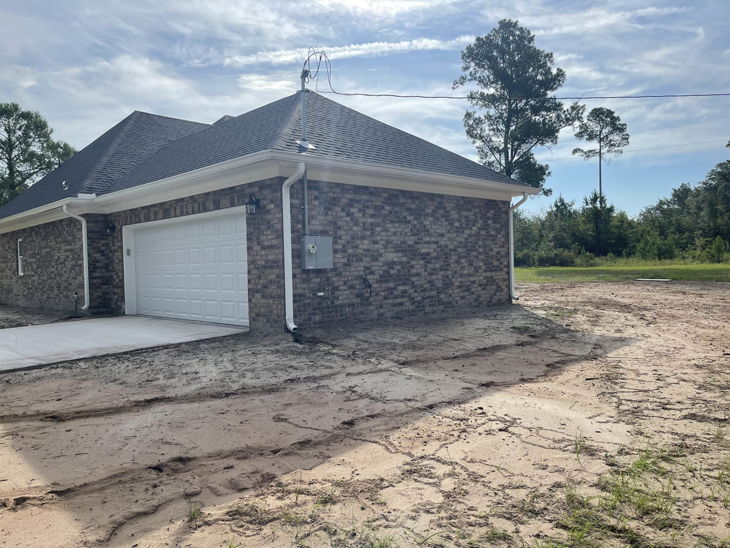 Red brick house with white garage door, brick trim, and concrete driveway with dirt and gravel, surrounded by open land and trees under a cloudy sky