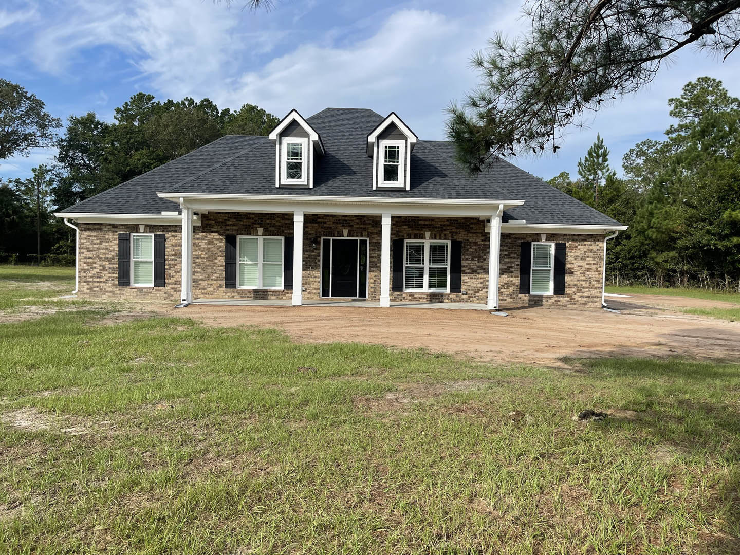Two-story house with white columns, black front door with glass panels, large green lawn, white-framed windows, and a dirt driveway bordered by grass.