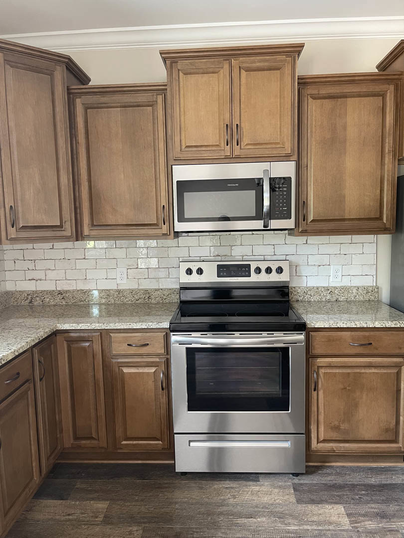Kitchen with natural wood cabinets, stainless microwave above stove, black cooktop with white burner lines, light wood flooring, white countertop, and partial view of a door.