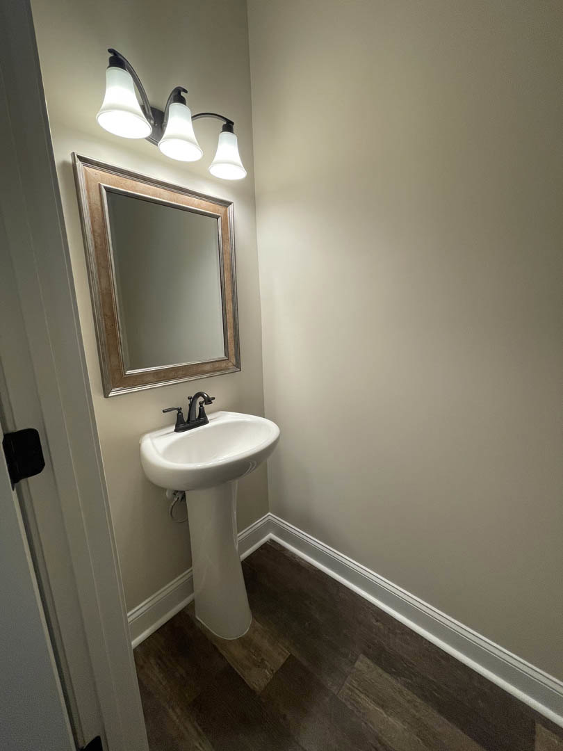 White pedestal sink with black faucet, three-light fixture above wall-mounted mirror, light tile floor and neutral wall in bathroom.