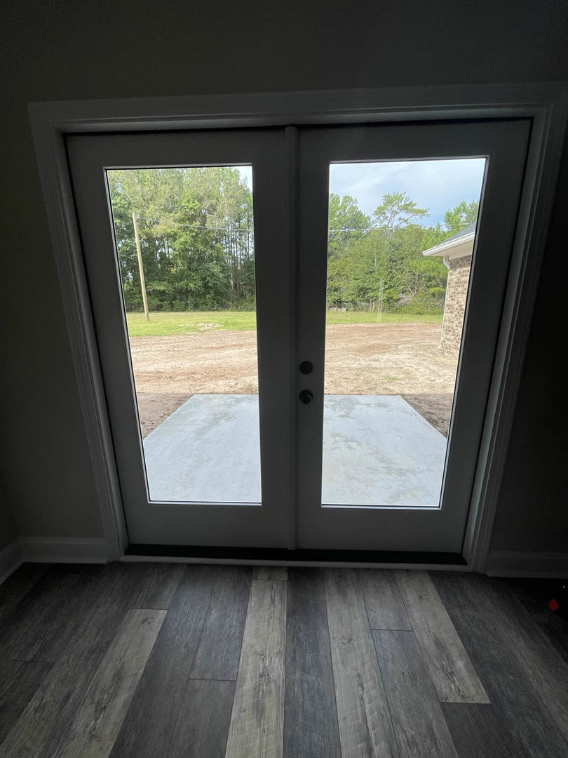 Glass door framed in wood, opening onto a landscaped yard with mature trees, sunlight streaming onto light hardwood flooring inside