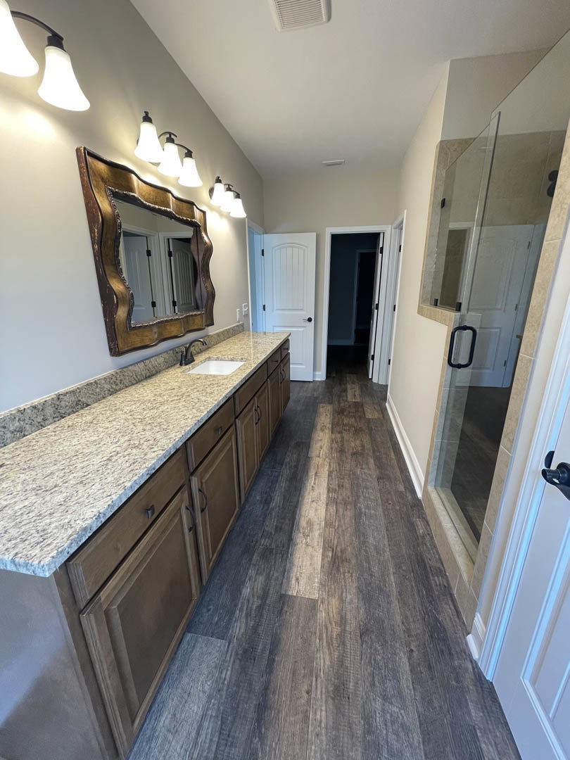 Bathroom featuring a marble countertop with undermount sink, large wall mirror, wood flooring, white door with black handle, and glass shower door.