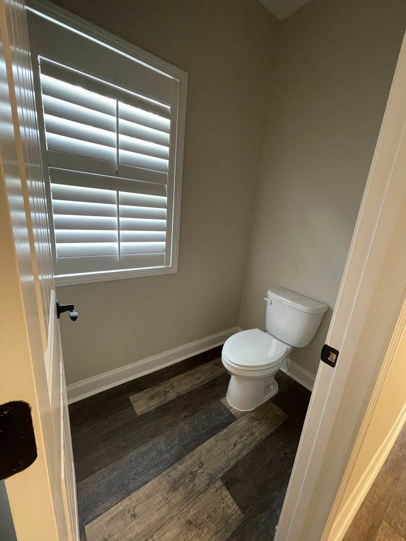 White toilet on wood floor beneath window with white blinds in bathroom with light tile walls