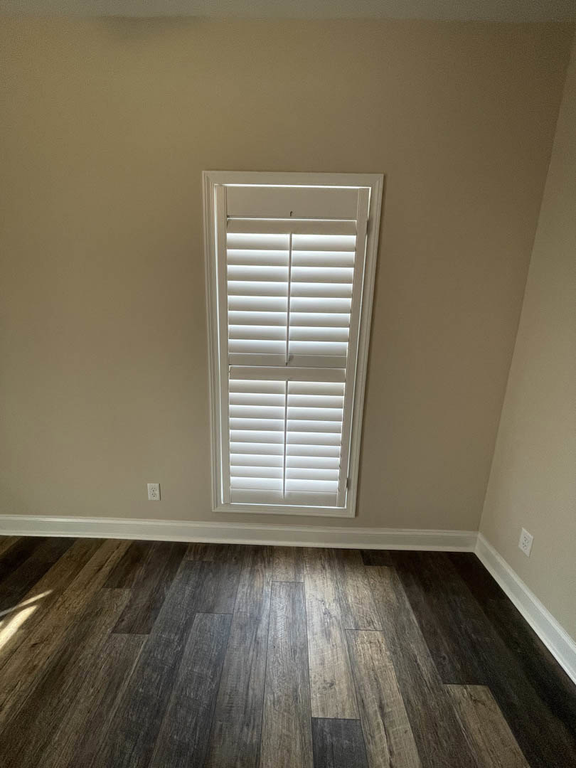 White window with closed blinds set in a plaster wall above dark wood laminate flooring and white baseboard