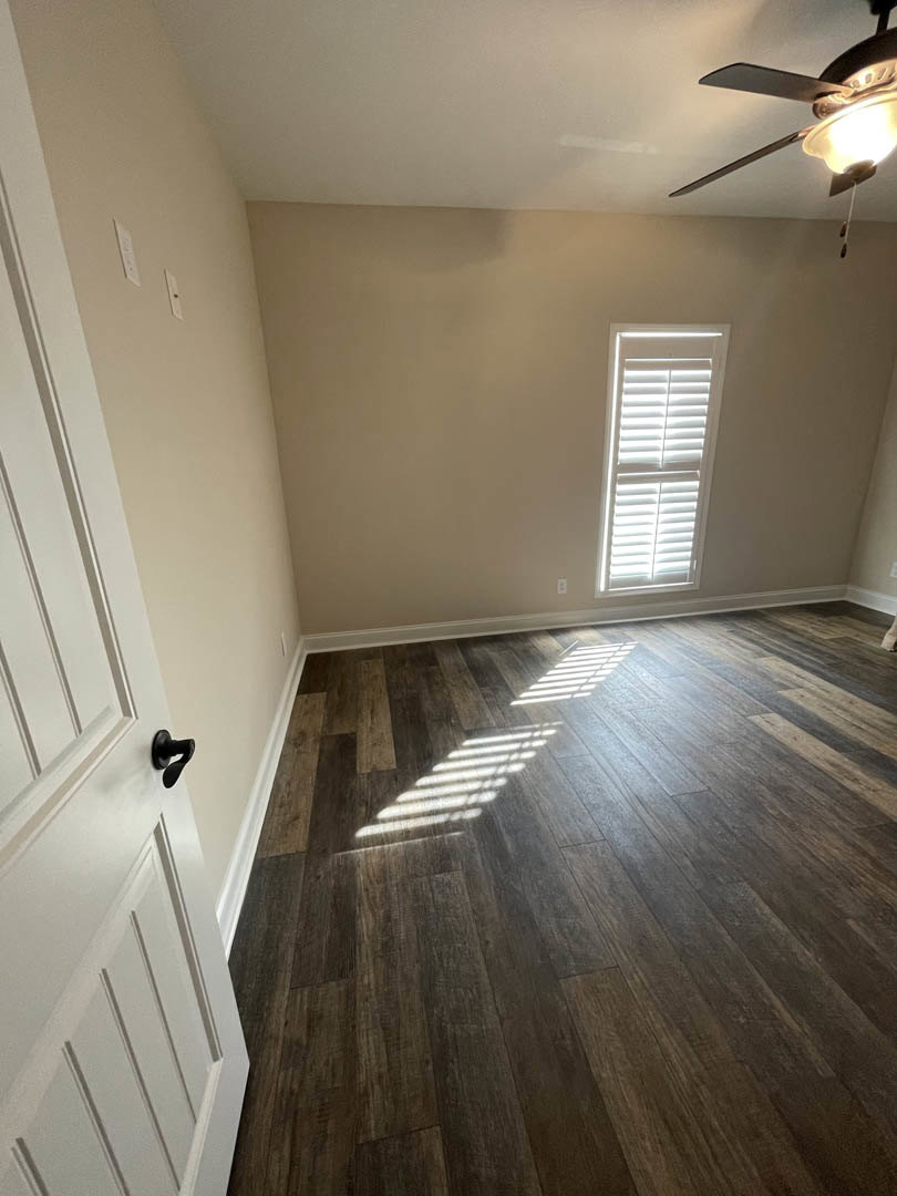 Wood flooring and ceiling fan in a bright room with large window, white walls, and sunlight filtering through blinds
