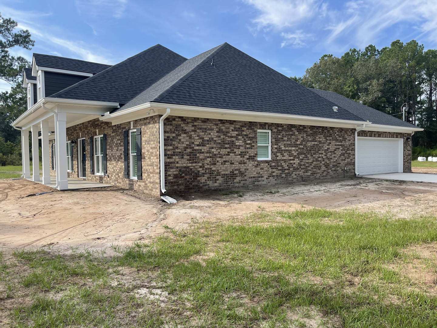 Brick house with white roof and white garage door, driveway bordered by green grass, windows with white frames and blinds, cloudy sky overhead, trees in background