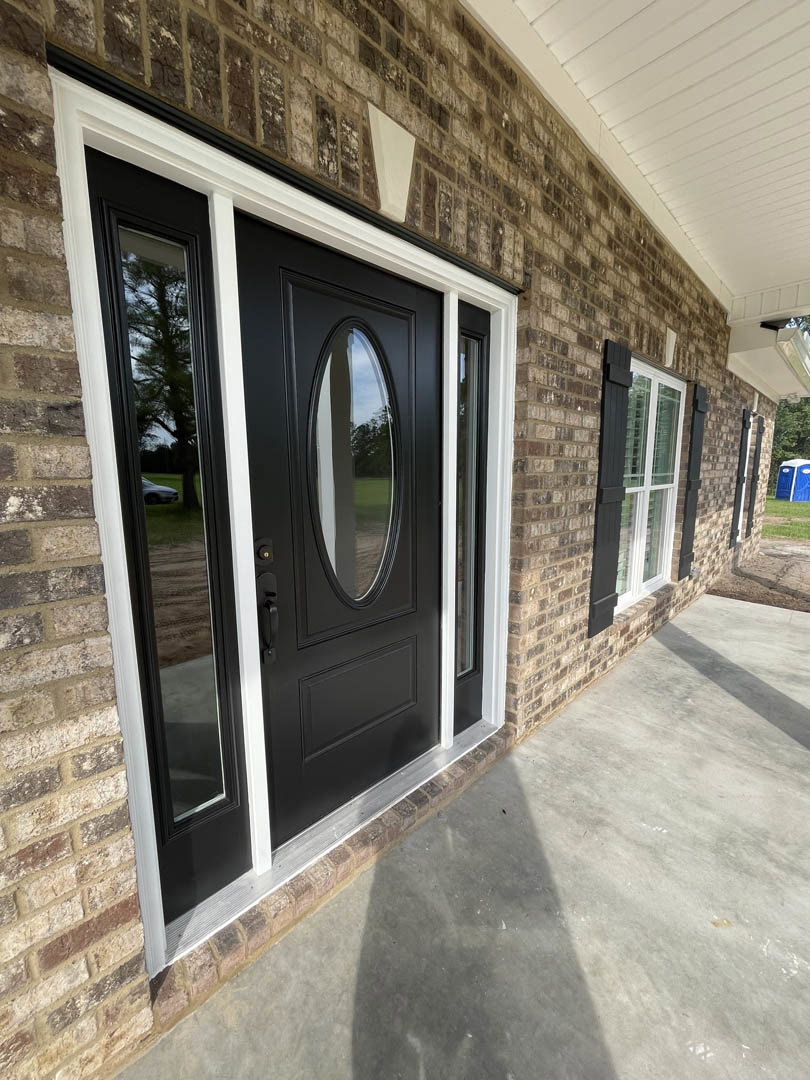 Black front door with oval glass window and silver hardware set in red brick wall, adjacent to window with white shutters reflecting green lawn