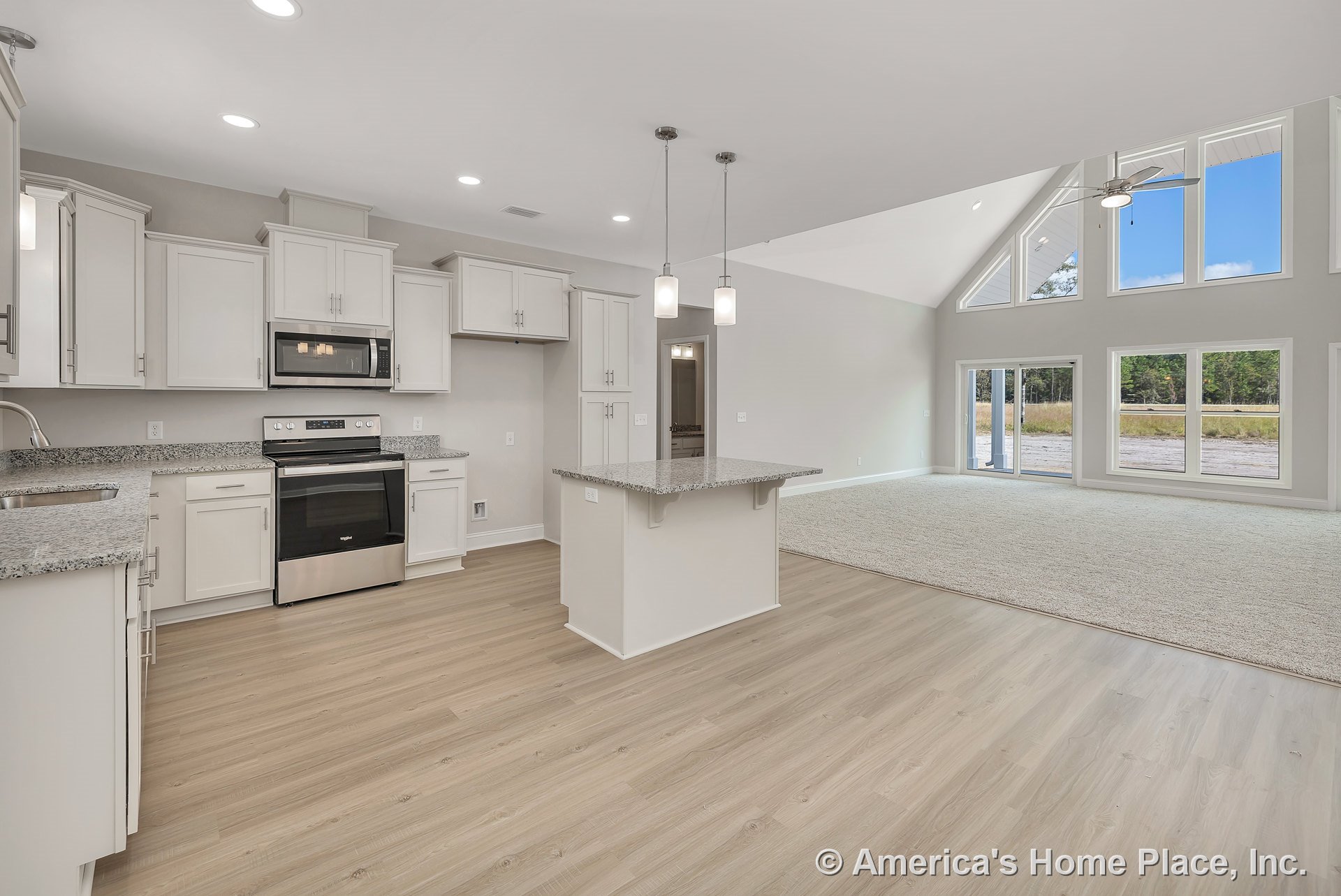 Open kitchen with white shaker cabinets, granite countertops, and a central island illuminated by pendant lights, seamlessly connected to a vaulted living area featuring expansive