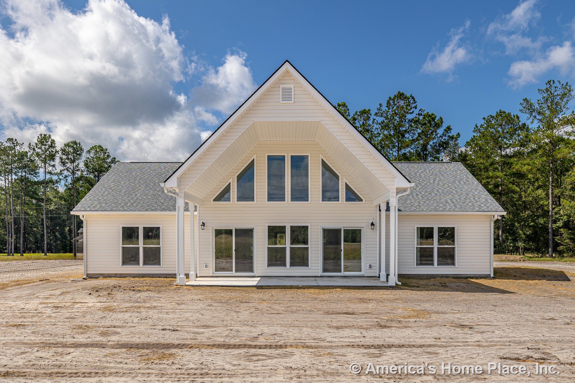 White exterior home with prominent gabled roof, vent detail, covered front porch, white-framed windows, and dirt driveway; trees and sky in background.