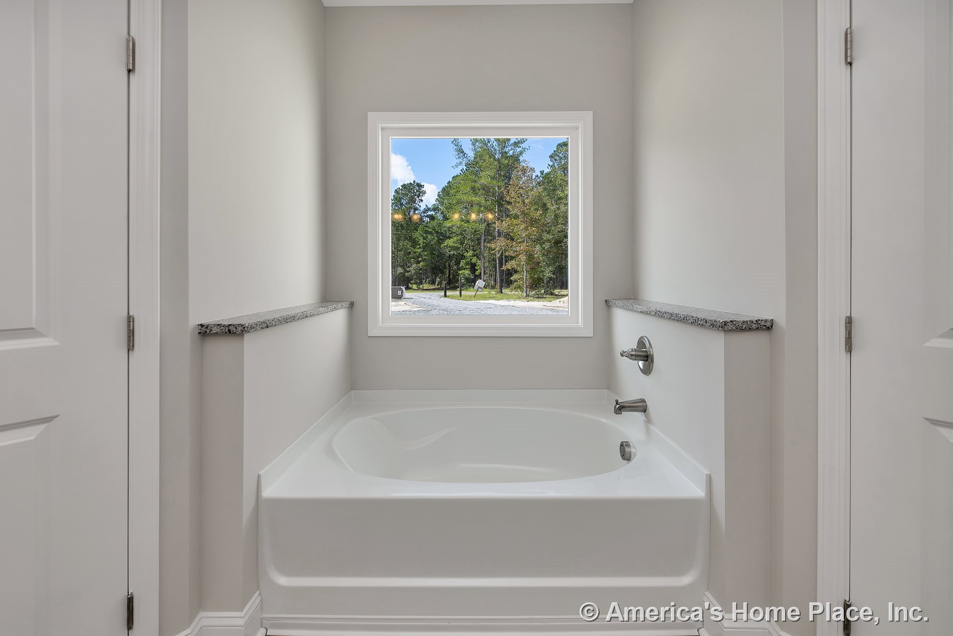 Built-in white soaking tub with chrome fixtures set beneath a large picture window, granite ledges surrounding the tub, neutral wall color, and trimmed window frame providing
