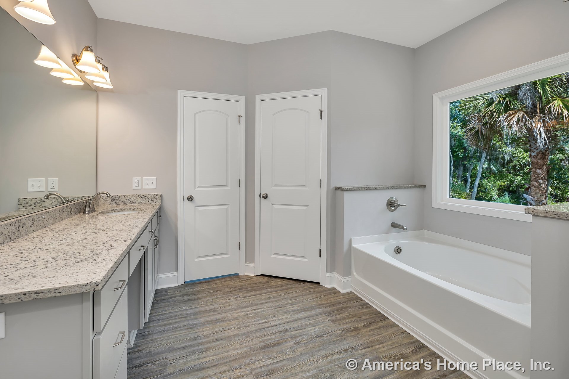 Soaking bathtub beside granite vanity countertop with large wall mirror, wood-look plank flooring, double pane window, dual doors, built-in overhead lighting, neutral paint, and