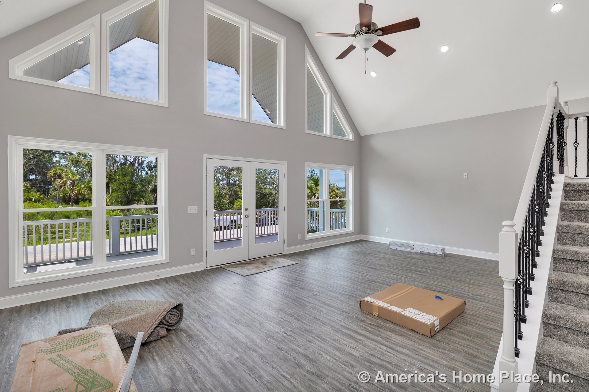 Vaulted ceiling living room with large triangular windows, double doors opening to covered porch, gray wood-look flooring, white trim, staircase with iron balusters, ceiling fan