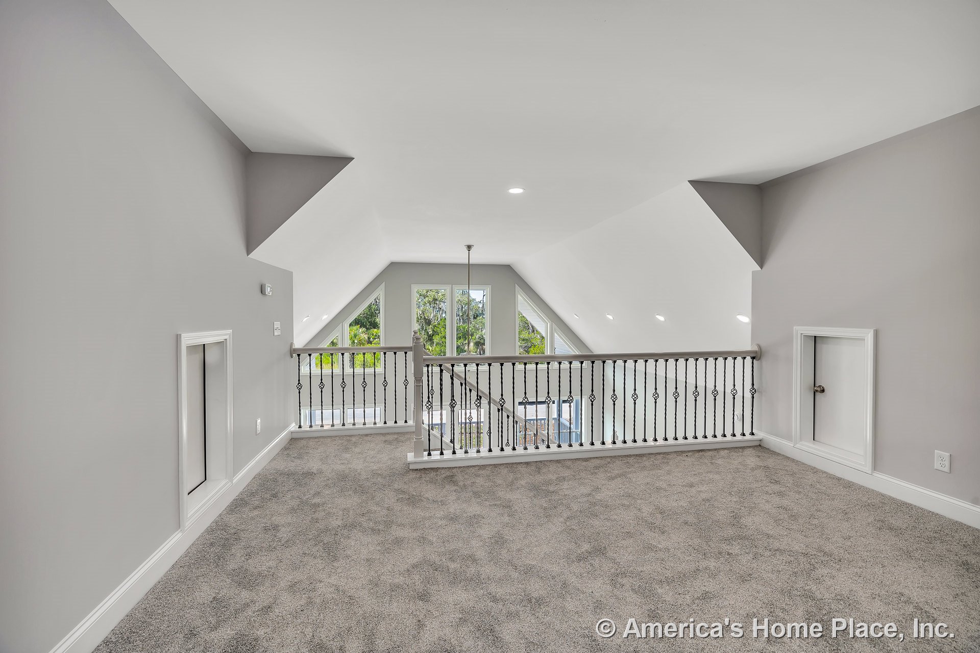 Carpeted loft with sloped white ceiling, decorative iron railing overlooking large triangular windows, built-in attic access doors, recessed ceiling lights, gray walls, and white