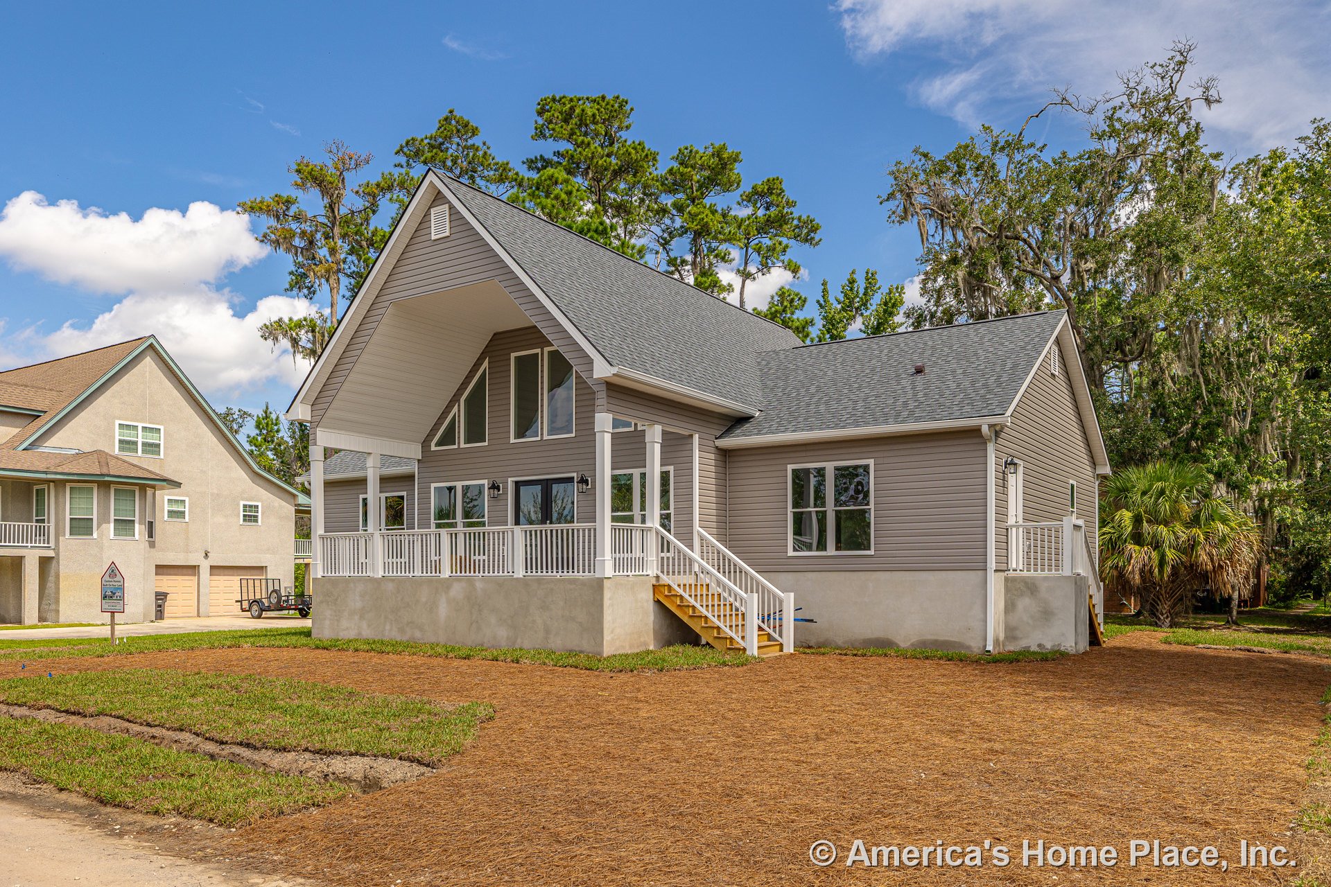 Large covered front porch with white railings, tall gable roof, multiple oversized windows, gray vinyl siding, exterior staircase, and concrete foundation.