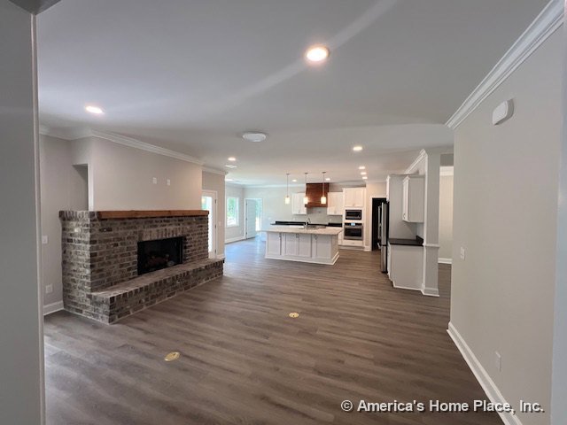 Open-concept living area with brick fireplace and wood mantel, recessed ceiling lights, white kitchen cabinetry, large kitchen island, wood-look plank flooring, crown molding trim